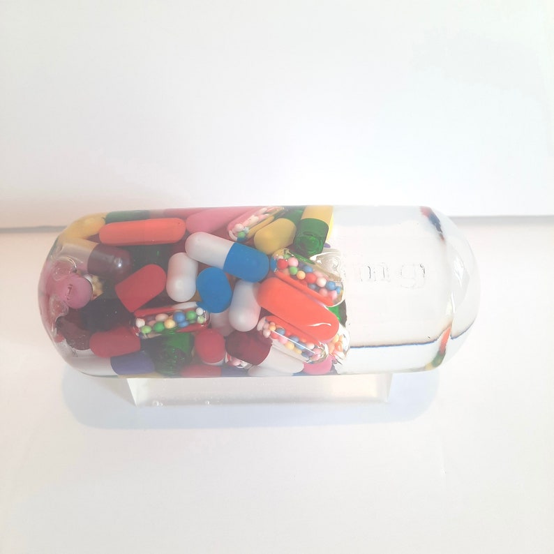 May include: A clear glass bottle filled with colourful pills and capsules. The bottle is on a white stand.