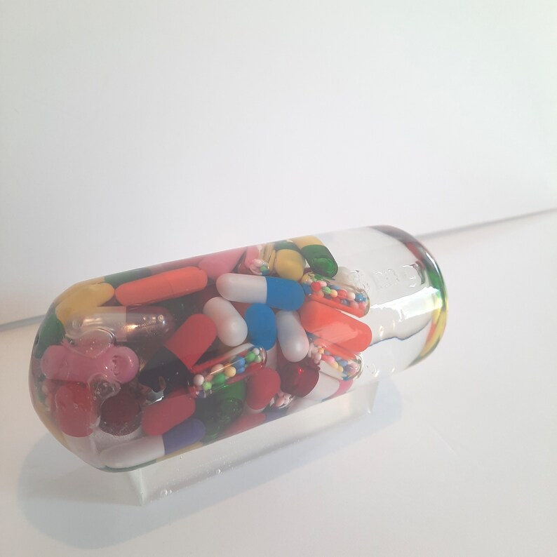 May include: A clear glass bottle filled with colourful pills and capsules. The bottle is on a white surface.