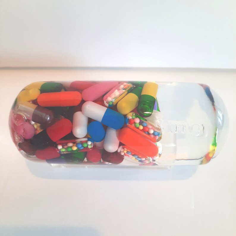May include: A clear glass capsule filled with colourful pills and capsules. The capsule is shaped like a pill and has a white label with the word "NOW" printed on it.