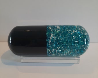 May include: A black and teal glitter filled capsule on a clear glass stand. The capsule is half black and half teal glitter.