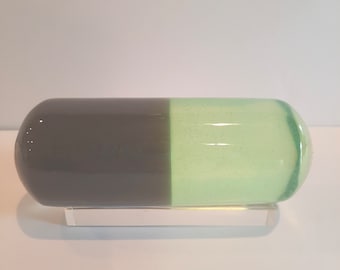 May include: A gray and green glass capsule on a clear acrylic stand. The capsule is shaped like a pill and has a smooth, rounded surface.