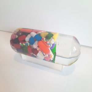 May include: A clear glass capsule filled with colourful pills, capsules, and sprinkles. The capsule is on a white surface.