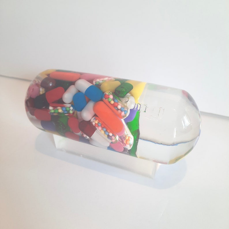 May include: A clear glass capsule filled with colourful pills, capsules, and sprinkles. The capsule is on a white surface.