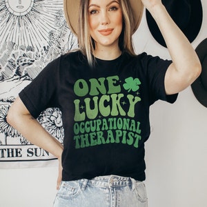 以下が含まれることがあります： クローバーが描かれた「One Lucky Occupational Therapist」という緑と白のグラフィックがプリントされた黒いTシャツです。
