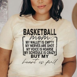 Könnte beinhalten: Ein hellbeiges T-Shirt mit einem schwarzen Aufdruck, der "Basketball Mom" mit einem Herzsymbol zeigt. Der Aufdruck enthält auch den Text "My wallet is empty, My nerves are shot, My voice is hoarse, My schedule is crazy, But my heart is full."