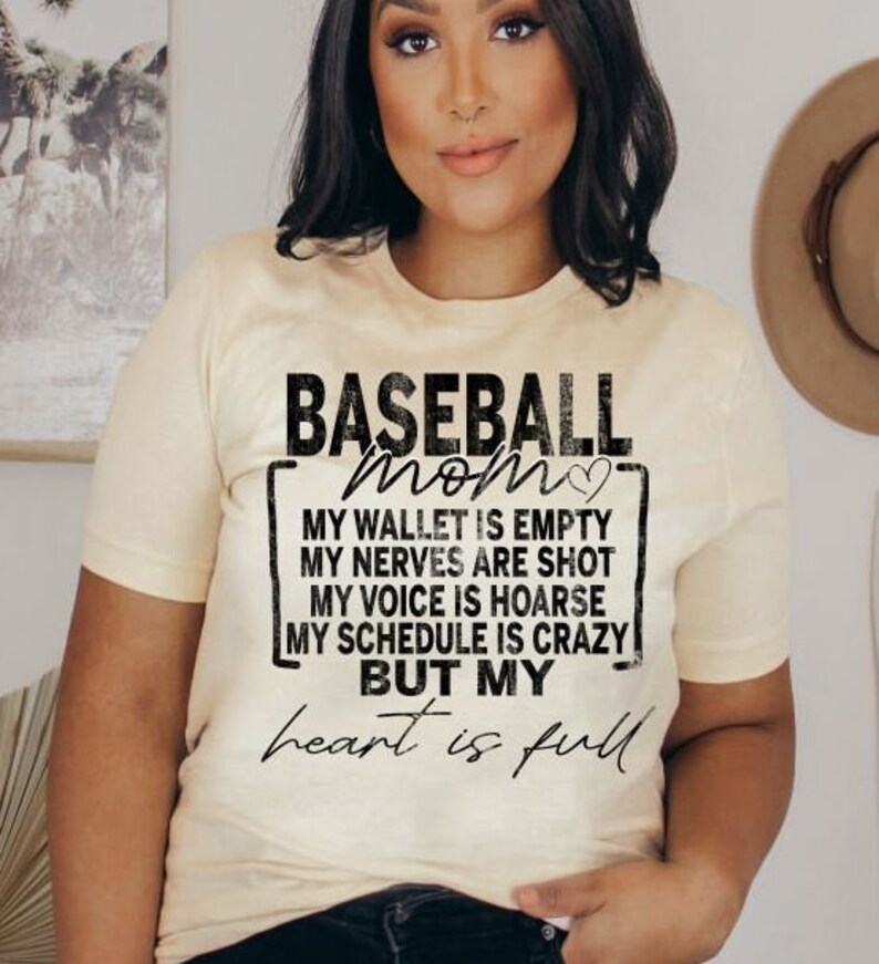 Peut inclure: Un t-shirt beige avec un motif noir qui dit "BASEBALL mom" avec un symbole de c&oelig;ur. Le motif comprend &eacute;galement le texte "MY WALLET IS EMPTY, MY NERVES ARE SHOT, MY VOICE IS HOARSE, MY SCHEDULE IS CRAZY BUT MY heart is full".