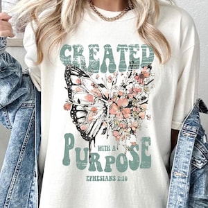 Peut inclure: T-shirt blanc avec un motif de papillon et de fleurs. Le texte "CREATED WITH A PURPOSE" est imprimé sur le t-shirt, ainsi que la référence biblique "EPHESIANS 2:10".