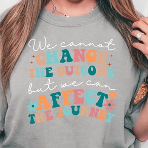 Puede incluir: Una camiseta gris con el texto "We cannot change the outcome but we can affect the journey" en letras de colores y estilizadas.
