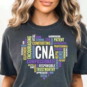 以下が含まれることがあります： 白い「CNA」の文字が入ったハート型のデザインの濃い灰色のTシャツ。「思いやり」、「親切」、「信頼できる」などの言葉が様々な色でデザインされており、看護師に関連する資質を強調しています。