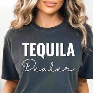 May include: A dark grey t-shirt with the text "TEQUILA Dealer" in white lettering. The text "Dealer" is written in a cursive font.