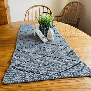 May include: A grey crocheted table runner with a diamond pattern on a wooden table. A small white wooden sign with the word "blessed" on it and a white ceramic bunny are on the table runner. A potted plant with green leaves and white flowers is in a black metal basket.