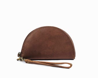May include: A semi-circular brown leather wristlet pouch featuring a smooth, matte finish. The accessory includes a matching brown leather wrist strap attached to a metal clasp and a zip closure that runs along the curved top edge of the pouch.