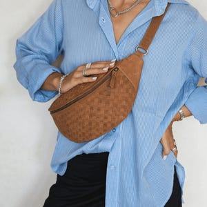 May include: A brown belt bag featuring a woven texture and a top zipper closure. The bag is worn across the chest with an adjustable strap attached to a silver-toned metal ring. The surface of the bag displays a consistent grid-like pattern.