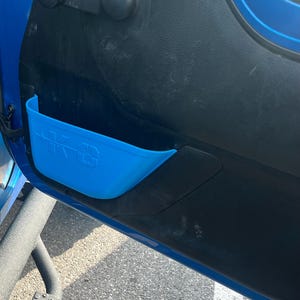 Può includere: Un pannello porta interno blu con una maniglia nera e una tasca portaoggetti blu. La tasca ha il testo "K&S" stampato sopra. Il pannello della porta fa parte dell'interno di un veicolo.