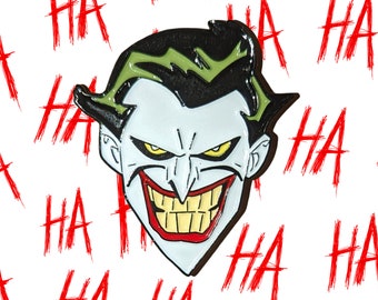 May include: A black and green enamel pin featuring a cartoon depiction of the Joker from Batman. The Joker has a wide, toothy grin and a green and black hair style. The background is white with red "HA" text repeated throughout.
