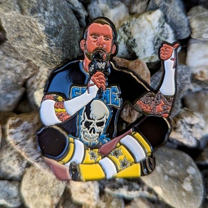 Könnte beinhalten: Eine schwarze und blaue Emaille-Anstecknadel, die einen sitzenden Wrestler mit einem Mikrofon in der einen Hand und einem Daumen nach oben in der anderen Hand zeigt. Der Wrestler trägt ein schwarzes Shirt mit einem Totenkopf-Motiv und dem Wort "C.M. Punk" darauf. Der Wrestler trägt auch eine gelbe und weiße gestreifte Hose.