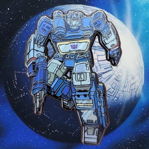 Puede incluir: Un pin de robot Transformers azul y plateado con un diseño detallado. El robot está en una pose de vuelo con los brazos extendidos. El pin está colocado sobre un fondo espacial azul y blanco.