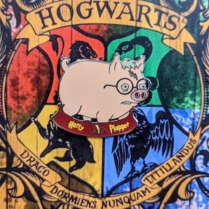 May include: A pink pig wearing glasses with a red and gold Hogwarts house crest pin. The crest says "Horty Plopper" and features a pair of glasses and a lightning bolt.