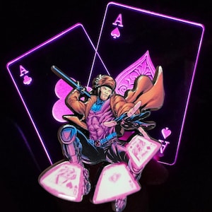 Pode incluir: Um broche de esmalte roxo e rosa com um personagem de banda desenhada, Gambit, numa pose dinâmica. Ele está segurando um cajado e em pé em cima de quatro cartas de jogar, todas as quais são ases de paus. O fundo é um par de cartas de jogar rosa brilhantes, ambas ases de paus.