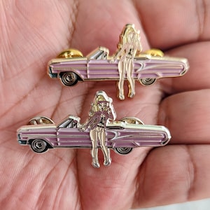 Può includere: Due spille smaltate raffiguranti un'auto convertibile rosa con una donna bionda in un abito rosa in piedi accanto. La donna indossa una blusa rosa e una camicia bianca sotto. L'auto ha un tetto bianco e una carrozzeria rosa. Le spille sono dorate.