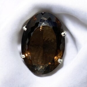 May include: A large, oval-shaped, brown gemstone ring set in silver. The gemstone is faceted and has a dark brown color.