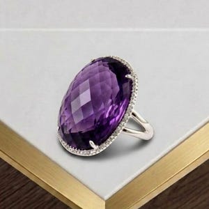 May include: A silver ring featuring a large, oval-shaped, faceted purple amethyst gemstone as the centerpiece. The stone is surrounded by a delicate halo of small, clear, round-cut stones set into the silver frame, which has a polished finish.