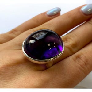 Pode incluir: Um anel de prata com uma grande ametista redonda em cabochão. A ametista é de cor roxa escura e está engastada em uma moldura de prata.