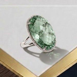 May include: A silver ring featuring a large, oval-cut, pale green gemstone as the centerpiece. The stone is surrounded by a delicate halo of tiny, clear, sparkling stones set into the silver frame, which connects to a smooth, polished silver band.