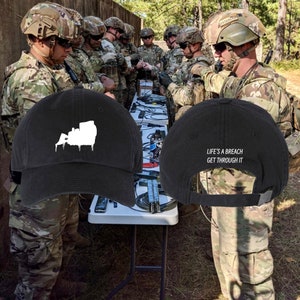 May include: Two black baseball caps with white embroidery. One cap features a silhouette of a bulldozer with dripping paint. The other cap has the text "Life's a breach get through it".