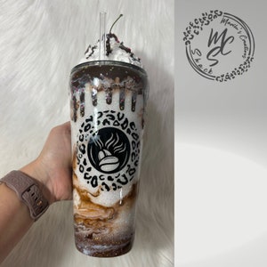 K&ouml;nnte beinhalten: Ein glitzernder wei&szlig;er Becher mit einer braun-wei&szlig; marmorierten Basis, einem zentralen schwarzen Kaffeebohnen-Symbol in einem Leopardenmuster-Kreis und einem k&uuml;nstlichen Schlagsahne-Topping mit Schokoladenguss, Streuseln und einer Kirsche.