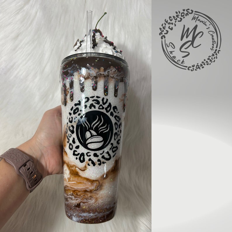 K&ouml;nnte beinhalten: Ein glitzernder wei&szlig;er Becher mit einer braun-wei&szlig; marmorierten Basis, einem zentralen schwarzen Kaffeebohnen-Symbol in einem Leopardenmuster-Kreis und einem k&uuml;nstlichen Schlagsahne-Topping mit Schokoladenguss, Streuseln und einer Kirsche.