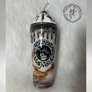 K&ouml;nnte beinhalten: Ein wei&szlig;er Glitzerbecher mit einem Kaffeebohnen-Logo, das von einem schwarzen Leopardenmuster-Kreis umgeben ist. Die Oberseite ist mit k&uuml;nstlicher Schlagsahne, Schokoladensauce, bunten Streuseln und einer Kirsche sowie einem Trinkhalm verziert.
