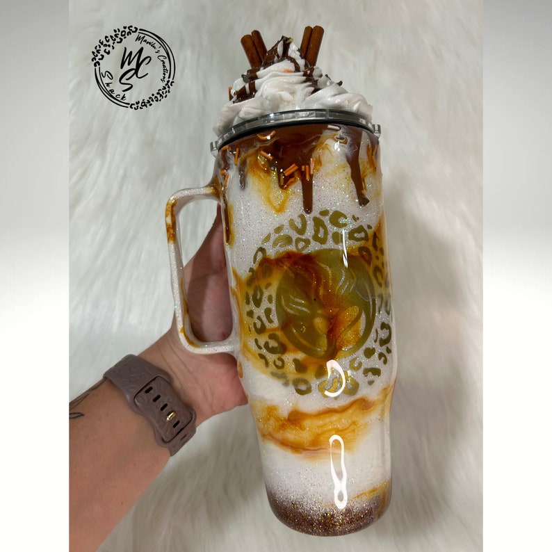 May include: A white and brown glitter tumbler with a leopard print design and a caramel drizzle. The tumbler has a white lid with whipped cream and cinnamon sticks on top.