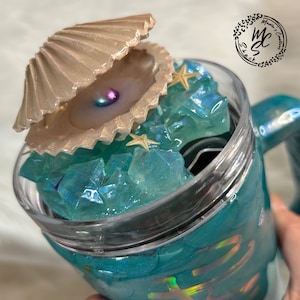 May include: A teal tumbler with a clear lid and a seashell charm. The lid is filled with blue and green iridescent crystals and a gold starfish. The tumbler has a holographic finish.