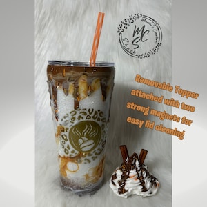 May include: A white and brown reusable tumbler with a removable lid and a straw. The tumbler has a brown and white swirl design with a gold coffee cup logo. The lid has a removable topper with a white whipped cream topping, chocolate drizzle, and two cinnamon sticks. The text "Removable Topper attached with two strong magnets for easy lid cleaning" is written in white on a black background.