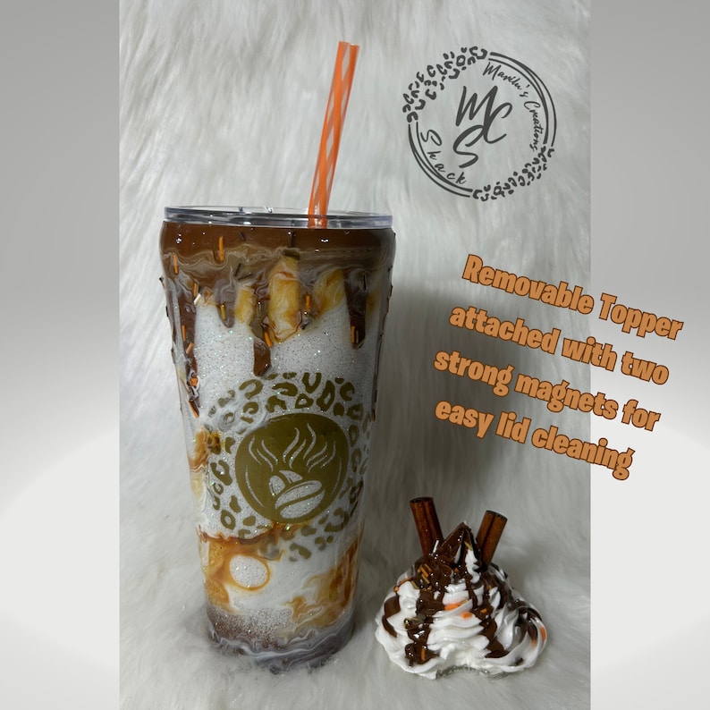 May include: A white and brown reusable tumbler with a removable lid and a straw. The tumbler has a brown and white swirl design with a gold coffee cup logo. The lid has a removable topper with a white whipped cream topping, chocolate drizzle, and two cinnamon sticks. The text "Removable Topper attached with two strong magnets for easy lid cleaning" is written in white on a black background.
