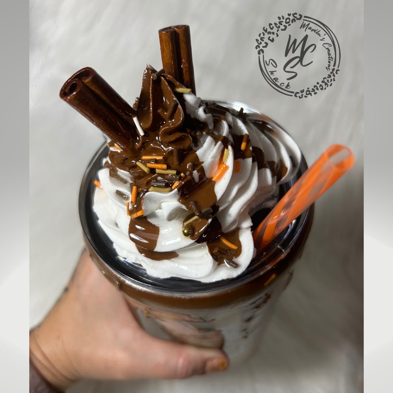 May include: A clear glass tumbler with a brown chocolate drink topped with whipped cream, chocolate sauce, sprinkles, and two cinnamon sticks. The tumbler has a lid and a straw.