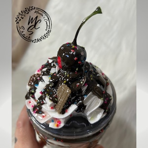 Puede incluir: Un vaso de plástico transparente lleno de un batido de chocolate cubierto con crema batida, salsa de chocolate, chispas y una cereza cubierta de chocolate.