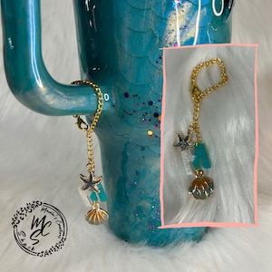 May include: A gold chain with a starfish, a seashell, and a turquoise charm attached. The chain is hanging from a teal tumbler with a mermaid tail design.