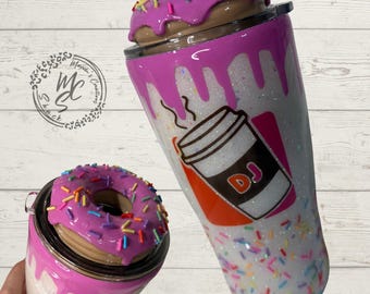 Könnte beinhalten: Ein weißer Glitzerbecher mit einem violetten Donut-Deckel mit bunten Streuseln, rosa Glasurtropfen und einer Grafik eines Kaffeebechers mit den Buchstaben DJ. Das Design enthält verstreute bunte Streusel auf dem unteren Teil des Bechers.