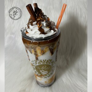 May include: A clear tumbler with a white lid and a brown and white design. The tumbler is filled with a white substance topped with whipped cream, chocolate sauce, and cinnamon sticks. The tumbler has a gold design with a coffee bean and flames.