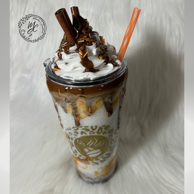 May include: A clear tumbler with a white lid and a brown and white design. The tumbler is filled with a white substance topped with whipped cream, chocolate sauce, and cinnamon sticks. The tumbler has a gold design with a coffee bean and flames.
