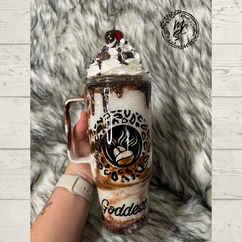 K&ouml;nnte beinhalten: Ein hoher Becher mit Henkel, wei&szlig;em Glitzer-Finish, einem schwarzen Leopardenmuster-Band und einem zentralen runden Logo mit Kaffeebohnen-Grafik. Die Oberseite ist mit k&uuml;nstlicher Schlagsahne, Schokoladenguss und einer Kirsche verziert.
