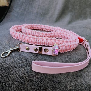 Puede incluir: Una correa para perro de color rosa claro que presenta una sección de paracord trenzado y un asa de correa plana y lisa. La correa incluye un mosquetón giratorio de metal plateado, tres remaches metálicos y un pequeño dije de esmalte colorido con forma de perro manchado.