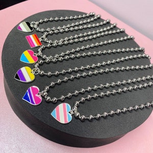 Op de afbeelding: Zeven zilveren kettingen met hartvormige hangers met verschillende pride-vlagontwerpen. De vlaggen omvatten de transgender-, bi-, pan-, non-binair- en genderqueer-vlaggen.