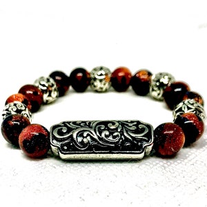 Puede incluir: Una pulsera de cuentas con cuentas rojas-marrones y plateadas alternas. La pulsera tiene un cierre plateado con un diseño ornamental y ondulado. Las cuentas rojo-marrones tienen un aspecto moteado y brillante.