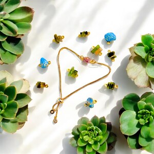 May include: A gold bracelet with a fine chain and a heart-shaped design, featuring colourful gemstone beads. Additional gemstone beads in green, blue, and pink are scattered around. The image is set against a white background with green succulents.