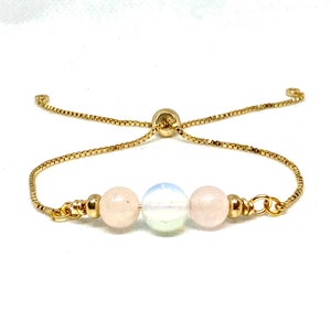 Könnte beinhalten: Verstellbares Armband in Goldoptik mit drei Edelsteinperlen. Das Armband hat eine filigrane Kette und einen Schiebeverschluss. Die Edelsteine umfassen zwei rosa Rosenquarzperlen und eine irisierende Mondsteinperle.