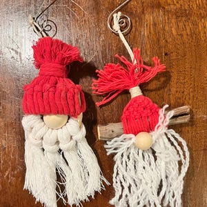 May include: Two handmade Christmas gnome ornaments. Each gnome has a red knitted hat, a wooden bead nose, and a white macrame beard. The ornaments are suspended by silver wire hangers. One gnome is attached to a small piece of wood.