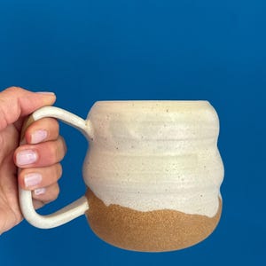 May include: Cream-colored ceramic mug with a brown base and a white handle. The mug has a textured, ridged design around the body. The background is a solid blue.